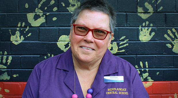 Joan Fraser is wearing a purple shirt, a name badge and red glasses. She is smiling. Behind her is a wall painted black and red with yellow paint hand prints from students.