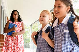 Two children wearing backpacks, looking excited in school environment with teacher in a pink dress watches on