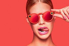 Female model wearing snapchat spectacles (glasses with cameras) against a peach background