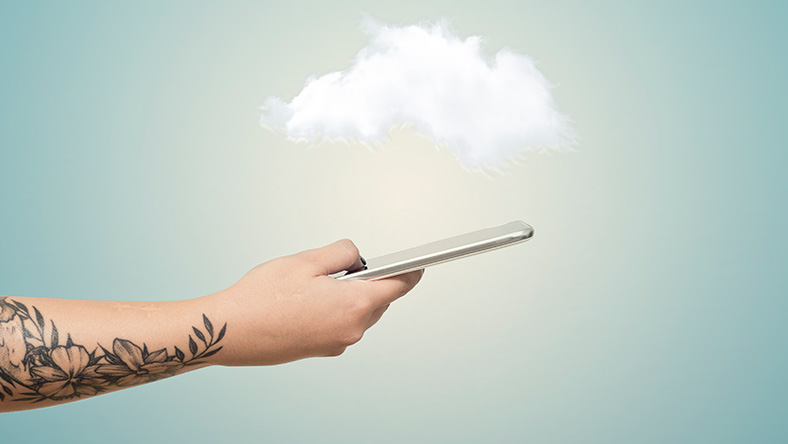 A person with a tattoo on forearm holds out a mobile phone while the foreground is made up of a single cloud and blue sky