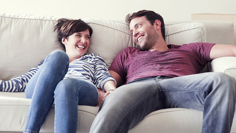 A happy pair of first home buyers lounge on the couch and smile at each other. 5 must read tips for first home buyers.