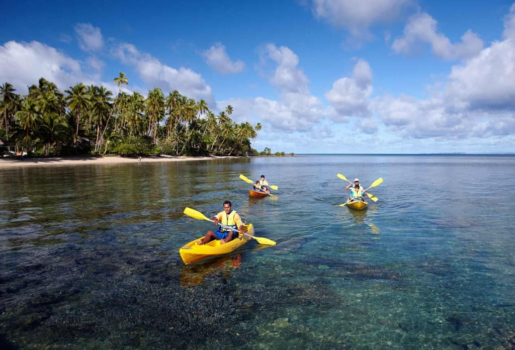 Three people kayaking in single kayaks across a coral reef near the shore of Fiji