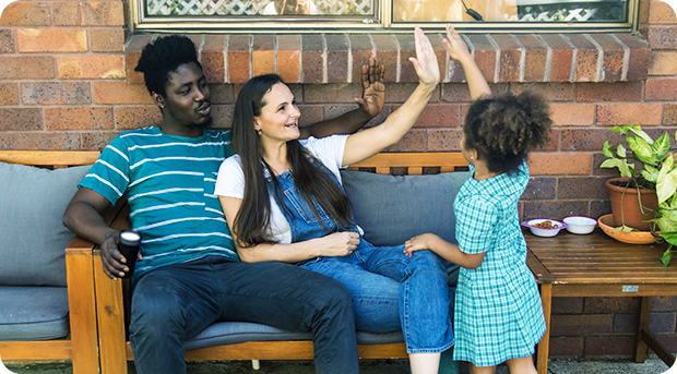 A family relax outside their new home. The mother and father are raising their hands to high-five their daughter.