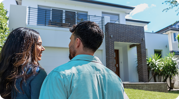 A couple stand out the front of their new home smiling at each other.